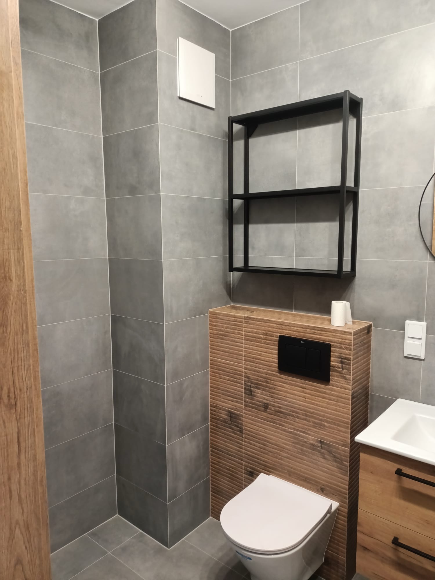 Nowoczesna łazienka z szarymi płytkami, drewnopodobną zabudową toalety, czarnym regałem i umywalką z drewnianą szafką.
