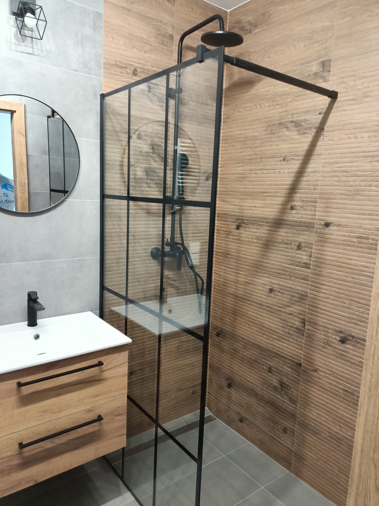 Nowoczesna łazienka z kabiną prysznicową typu walk-in z czarnymi profilami, umywalką nablatową na drewnianej szafce z czarnymi uchwytami i okrągłym lustrem z drewnianą ramą.
