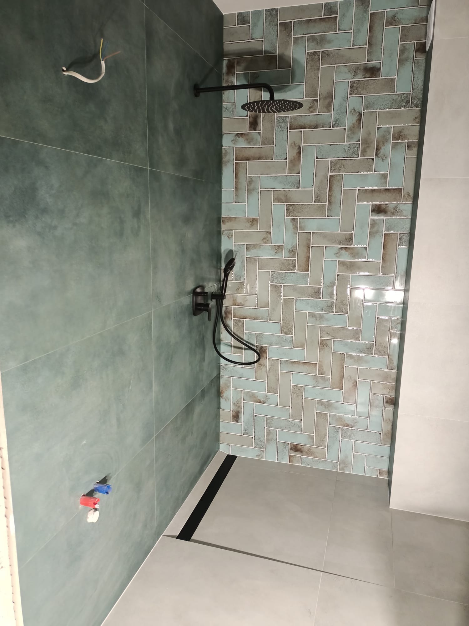 Wykończona kabina prysznicowa z zielonymi i szarymi płytkami ułożonymi w jodełkę, czarną armaturą i odpływem liniowym, widoczne wystające kable i rurki instalacji wodnej.