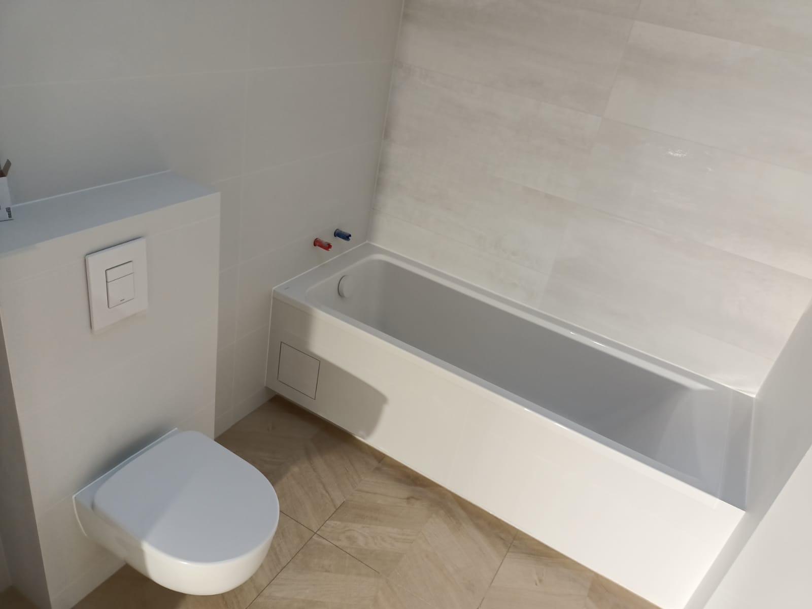 Minimalistyczna łazienka z białą zabudową WC i wanną, wykończona jasnymi płytkami imitującymi drewno na podłodze i szarymi, subtelnie wzorzystymi płytkami na ścianach.