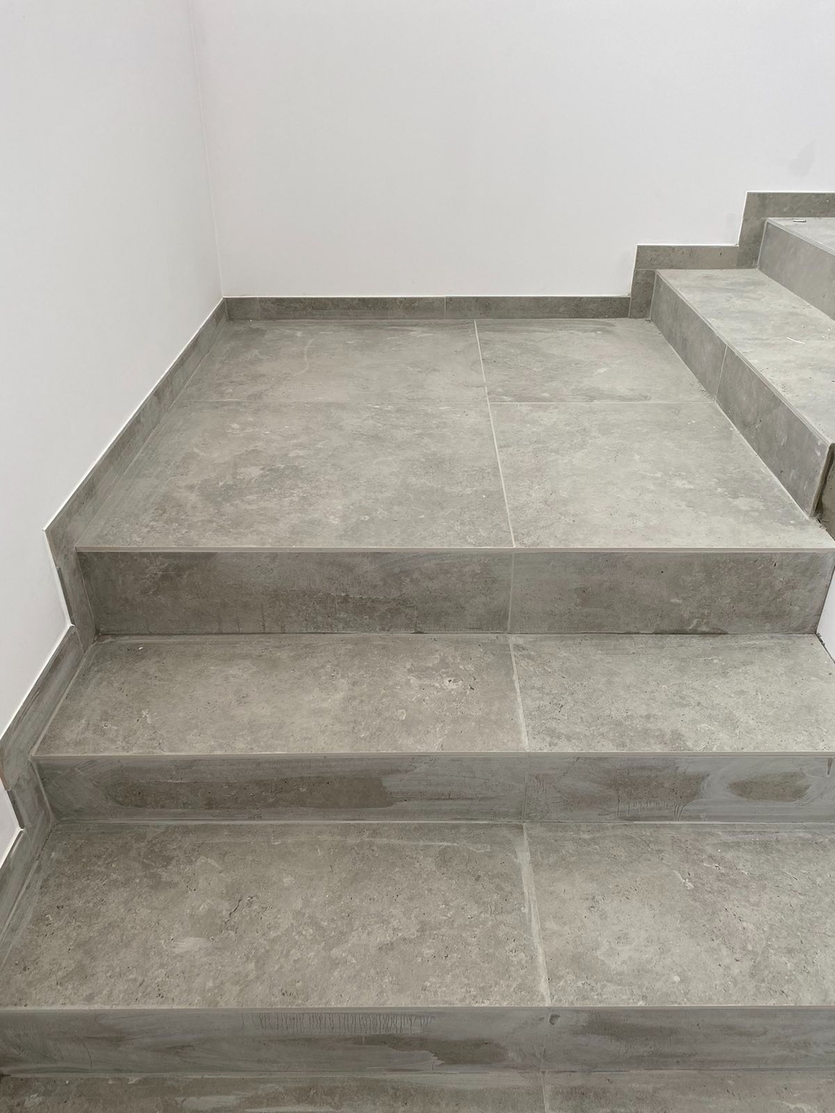 Wykończone schody wewnętrzne wyłożone szarymi płytkami imitującymi beton, z widocznymi fugami i białym narożnikiem ściany.