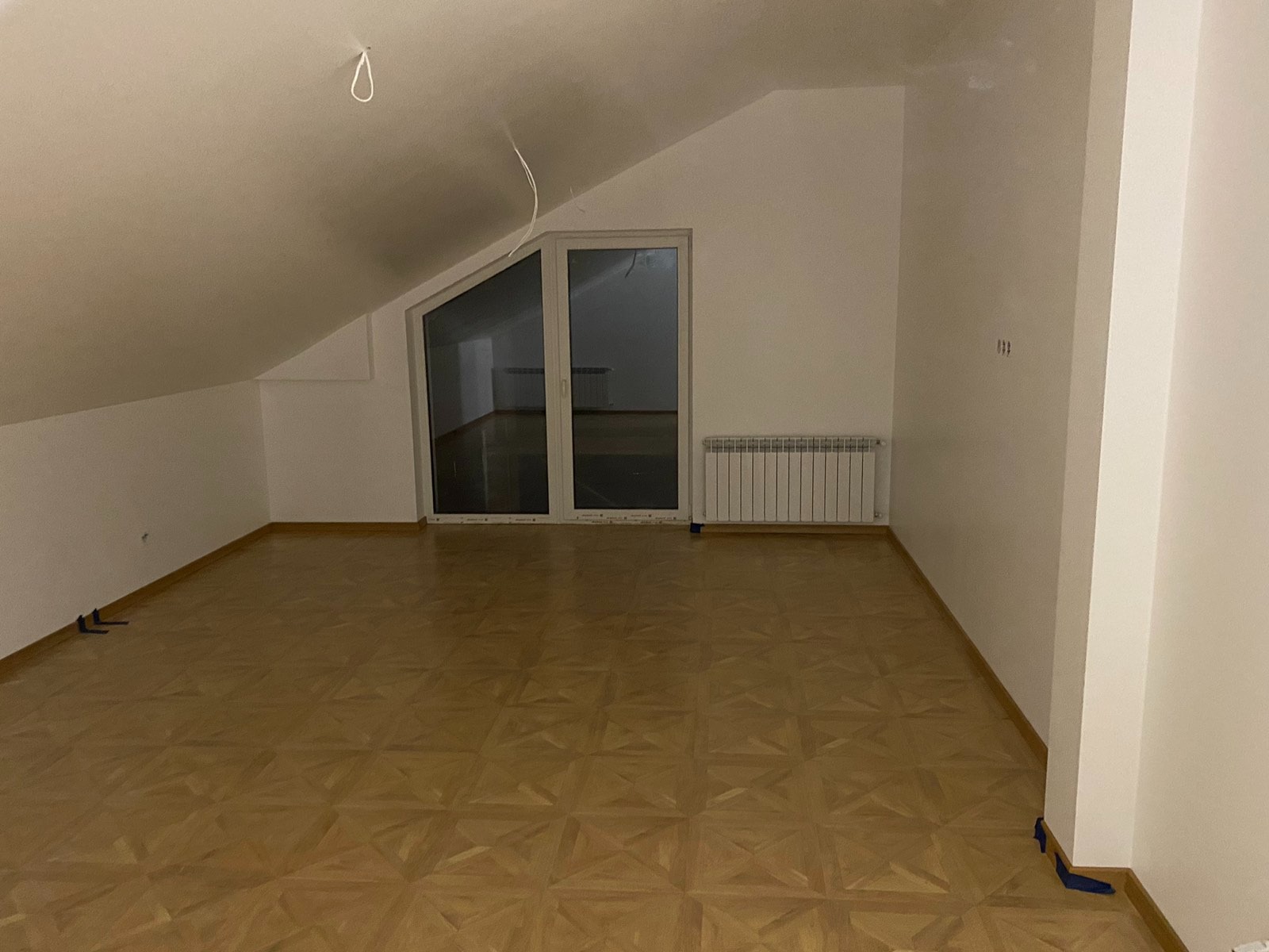 Puste poddasze ze skośnym sufitem, parkietem w jodełkę, grzejnikiem i oknem balkonowym przed remontem, widoczne kable wystające z sufitu.
