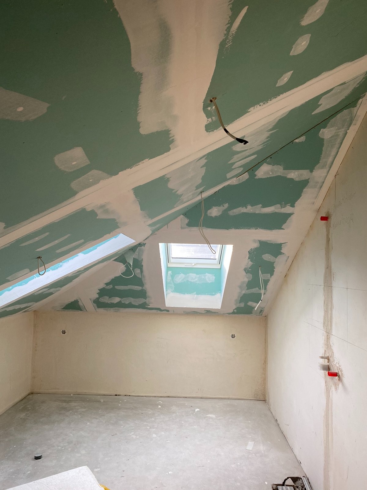 Poddasze w trakcie prac wykończeniowych: płyty gipsowo-kartonowe na skosach z widocznymi łączeniami pokrytymi masą szpachlową, okno dachowe, instalacje elektryczne wystające ze ścian, ślady po...