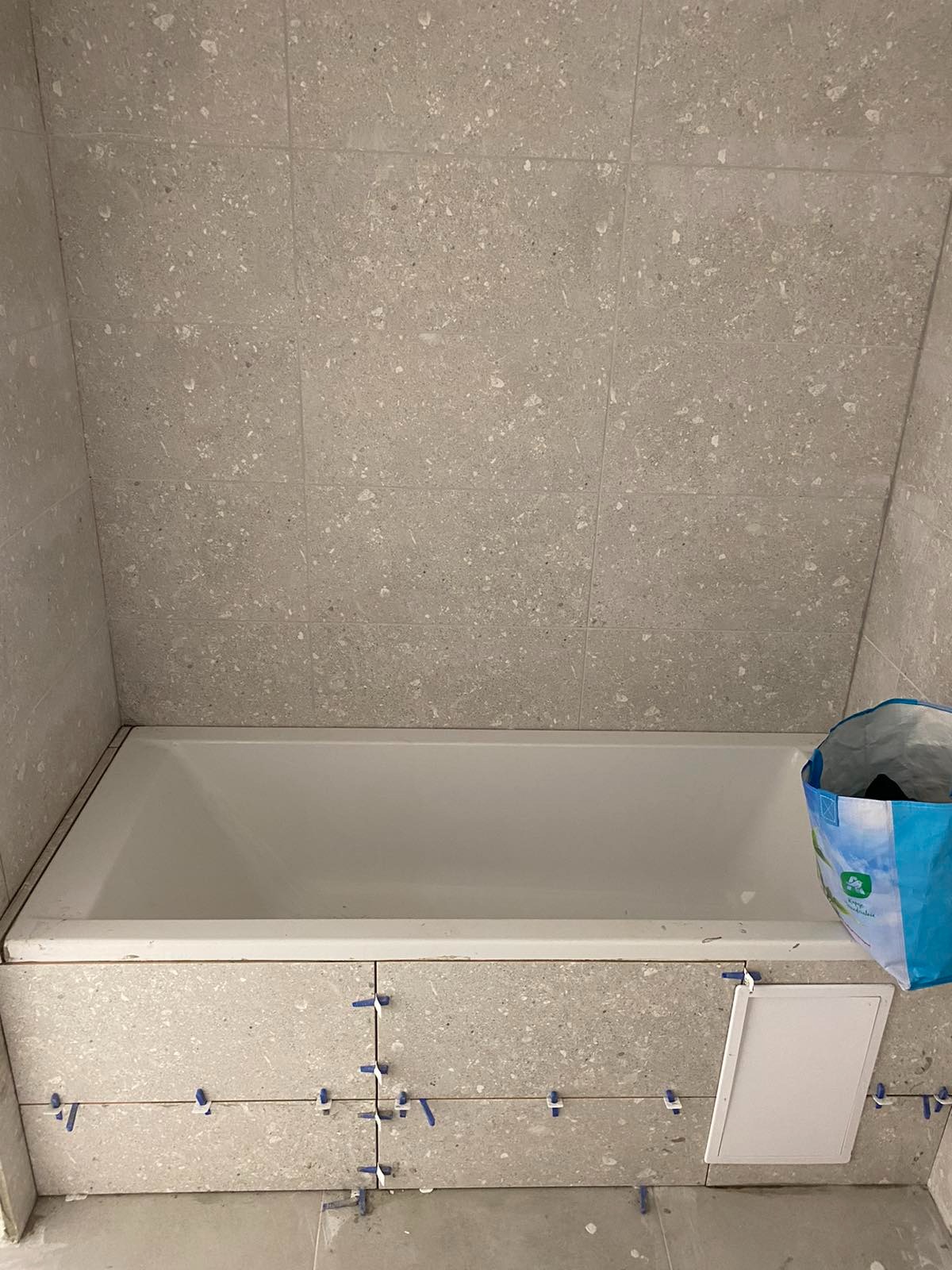 Biała wanna obudowana szarymi płytkami imitującymi kamień, widoczne krzyżyki dystansowe, niebieska torba na śmieci w rogu, mała biała klapka rewizyjna.