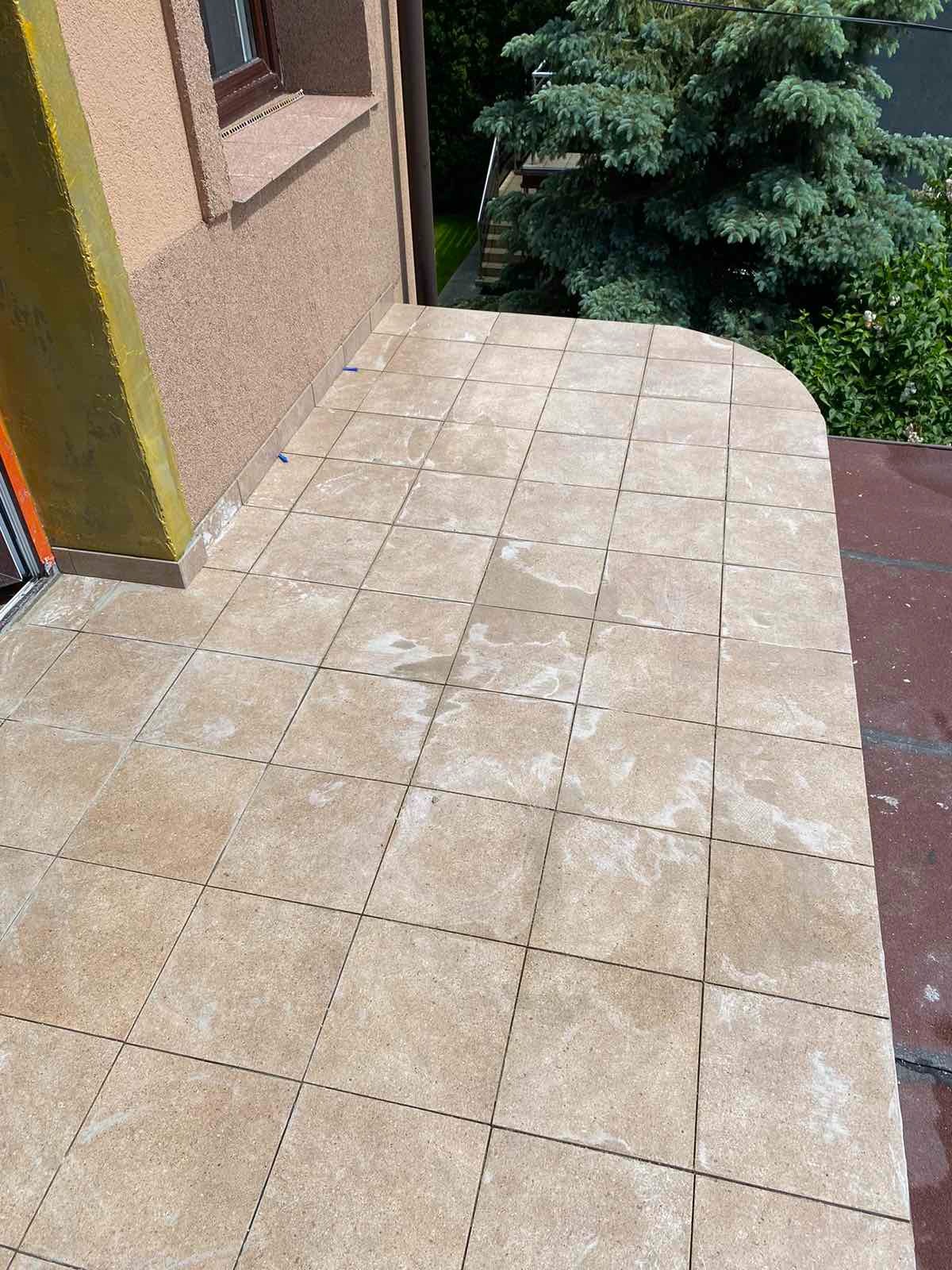 Wyłożony płytkami taras z widocznymi śladami białego cementu, fragment ocieplonej ściany budynku z oknem i zielone drzewo w tle.