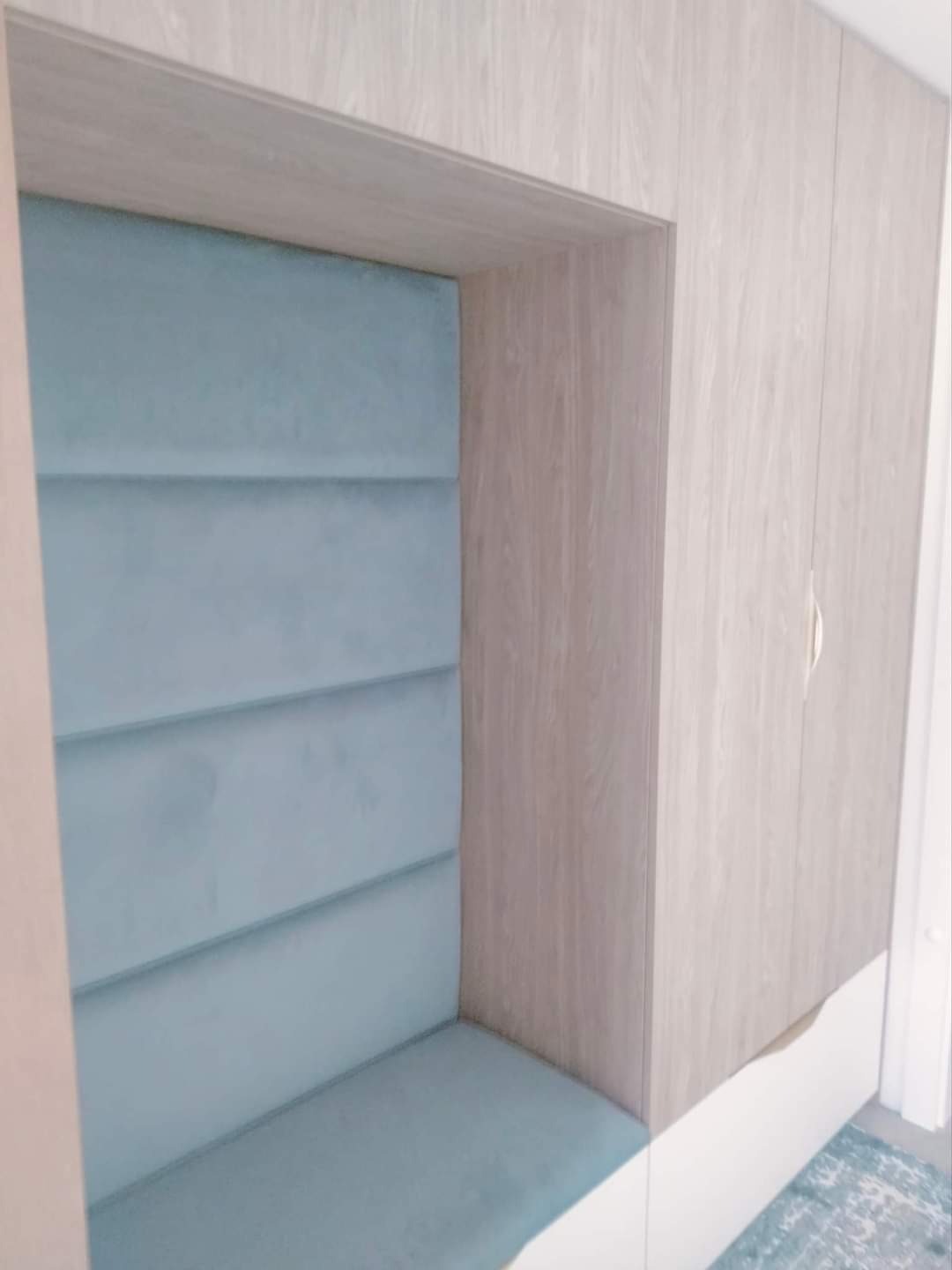 Wnęka z siedziskiem i tapicerowanym oparciem w odcieniach błękitu, obudowana drewnopodobną płytą meblową.