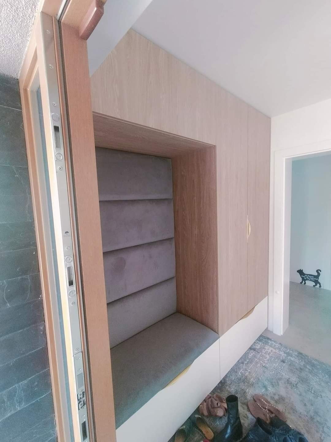 Wnęka z tapicerowanym siedziskiem i oparciem w zabudowie z drewnopodobnej płyty, widoczne buty na podłodze i dekoracja w kształcie kota w tle.