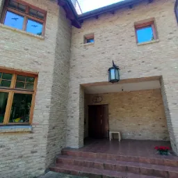 Dwupiętrowy dom z jasnej cegły klinkierowej z drewnianymi oknami, widok na wejście z zewnątrz, z małym stołkiem i kwiatami na schodach.