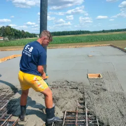 Pracownik w żółtych spodenkach wylewa beton na zbrojenie fundamentu domu, widoczne szalunki i otwarty teren w tle.