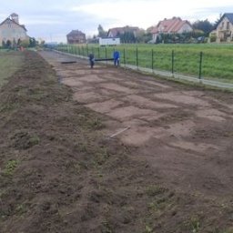 Niko-Bud - Przygotowany teren pod trawnik z widocznymi śladami po wyrównywaniu, w tle zabudowa jednorodzinna i zielone ogrodzenie z siatki.
