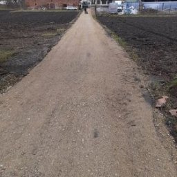 Niko-Bud - Wyrównany teren pod budowę drogi lub chodnika, widoczne świeżo rozplantowane podłoże z drobnym kruszywem, w tle zabudowania wiejskie i zaorane pole, pochmurne niebo.