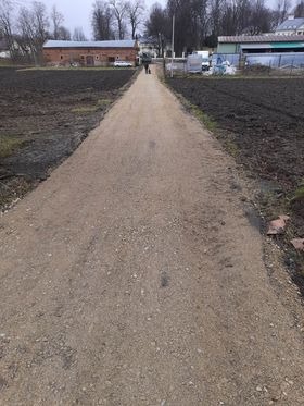 Wyrównany teren pod budowę drogi lub chodnika, widoczne świeżo rozplantowane podłoże z drobnym kruszywem, w tle zabudowania wiejskie i zaorane pole, pochmurne niebo.