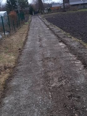 Przygotowany teren pod budowę drogi dojazdowej, widoczne wyrównane brzegi i ubita nawierzchnia, w tle pole uprawne i budynek gospodarczy.