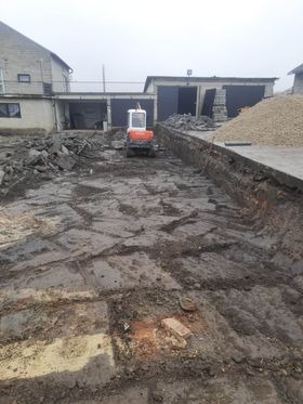 Mini koparka na placu budowy, przygotowany wykop pod fundamenty, widoczne ślady pracy, gruz i ziemia, w tle budynki gospodarcze i sterta kruszywa.