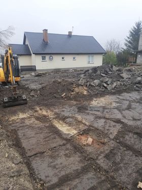 Żółta koparka na rozkopanym, błotnistym terenie przed domem z ciemnym dachem, widoczne ślady po płytach chodnikowych i gruz.