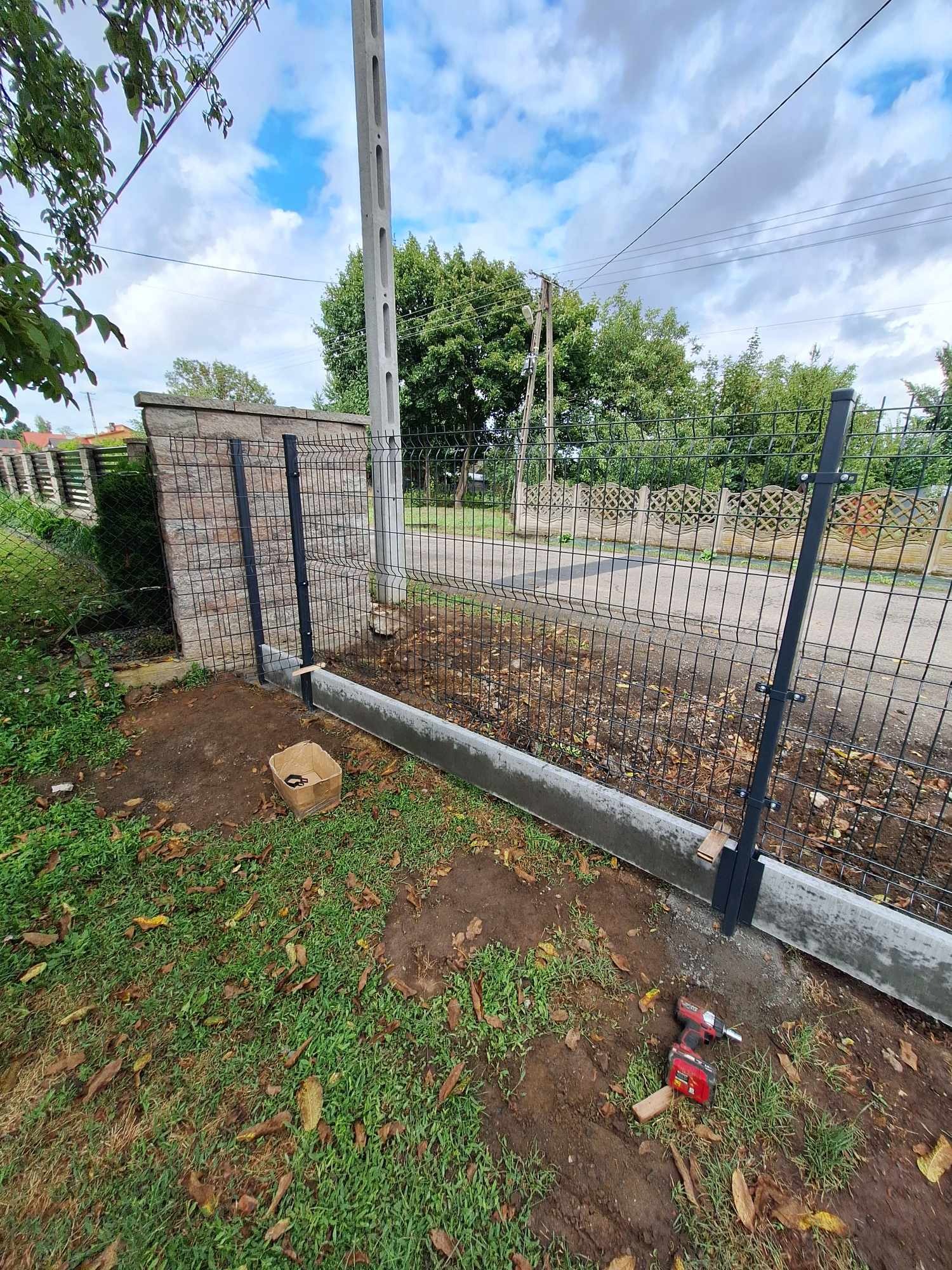 Montaż czarnych słupków ogrodzeniowych na betonowej podmurówce, widoczna wkrętarka akumulatorowa i kartonowe pudełko, w tle częściowo ukończone przęsła z siatki zgrzewanej.
