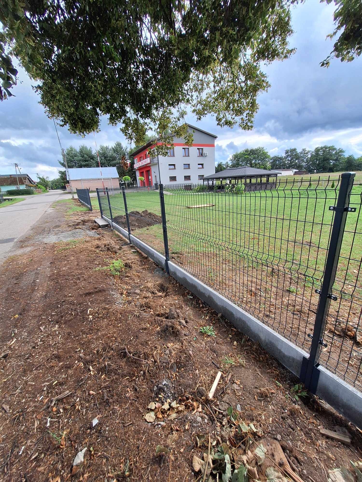 Nowo postawione, czarne ogrodzenie panelowe z podmurówką betonową, oddzielające posesję z budynkiem w tle od drogi. Świeżo usypana ziemia wzdłuż ogrodzenia.