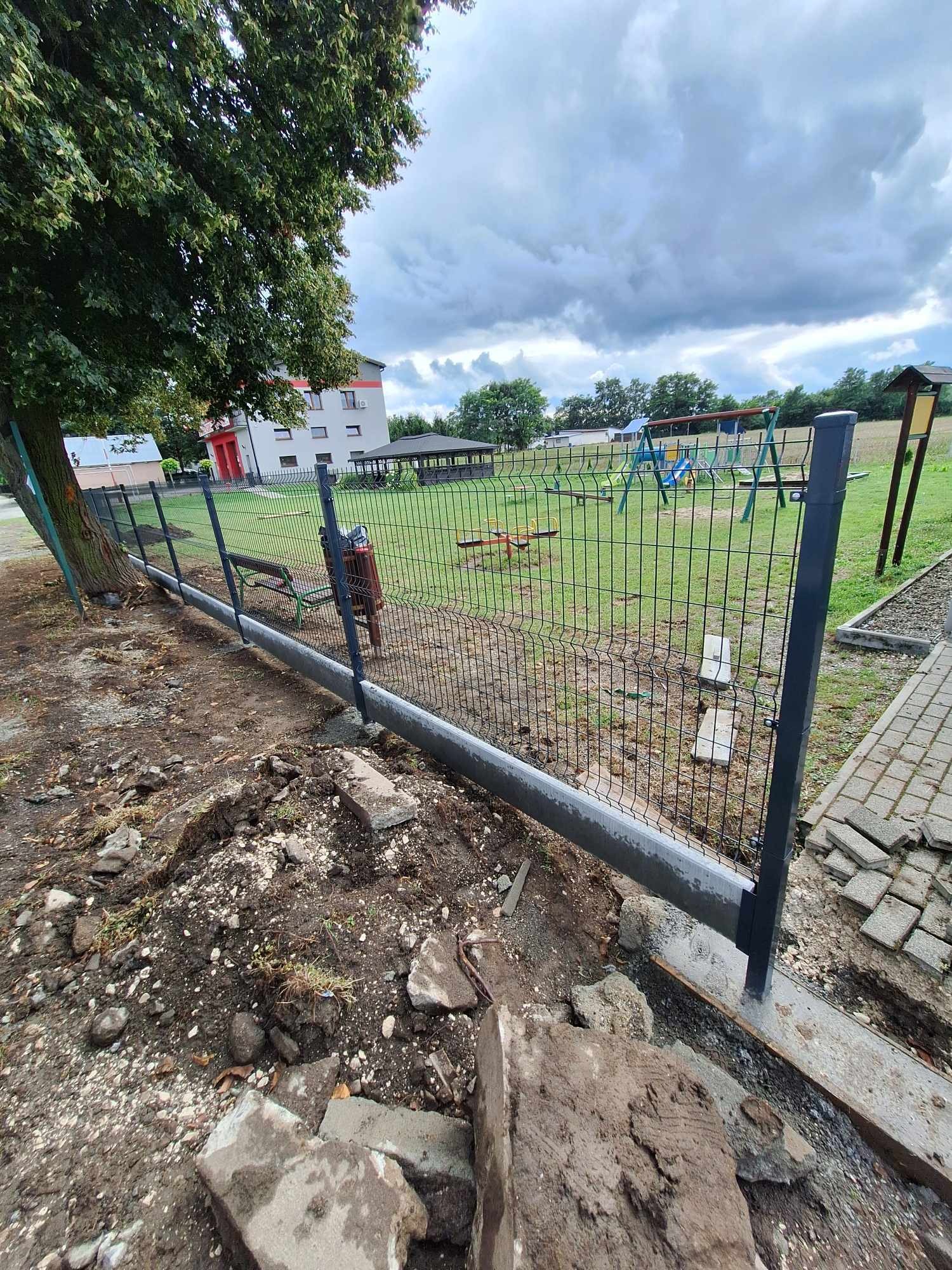 Montaż ogrodzenia panelowego z podmurówką betonową przy placu zabaw, widoczne słupki i siatka, ziemia rozkopana w trakcie prac.