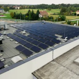 Widok z góry na dużą instalację paneli słonecznych na płaskim dachu budynku przemysłowego, z widocznymi elementami wentylacji i otaczającym krajobrazem.
