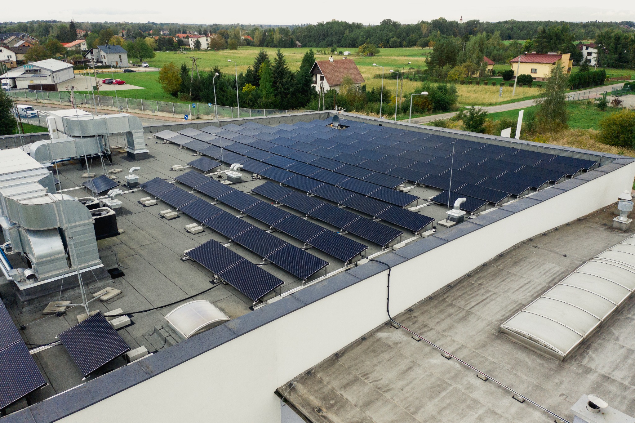 Widok z góry na dużą instalację paneli słonecznych na płaskim dachu budynku przemysłowego, z widocznymi elementami wentylacji i otaczającym krajobrazem.