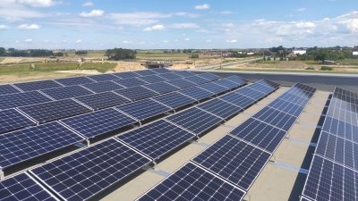 Rozległa instalacja paneli słonecznych na dachu budynku, widok z góry, w tle krajobraz z zabudowaniami i błękitne niebo z chmurami.