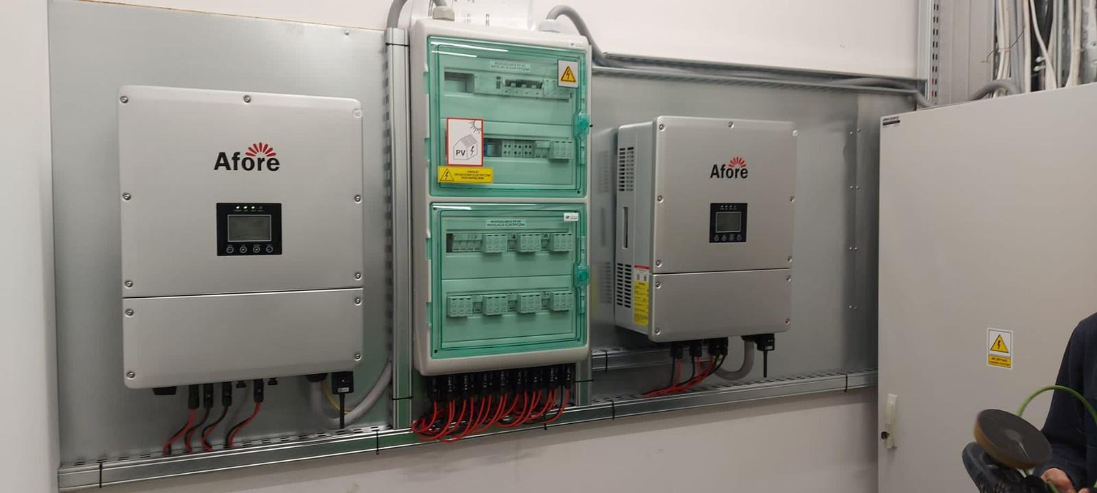 Dwa srebrne inwertery marki Afore zamontowane obok rozdzielnicy elektrycznej z zieloną obudową na metalowej szynie w pomieszczeniu technicznym.