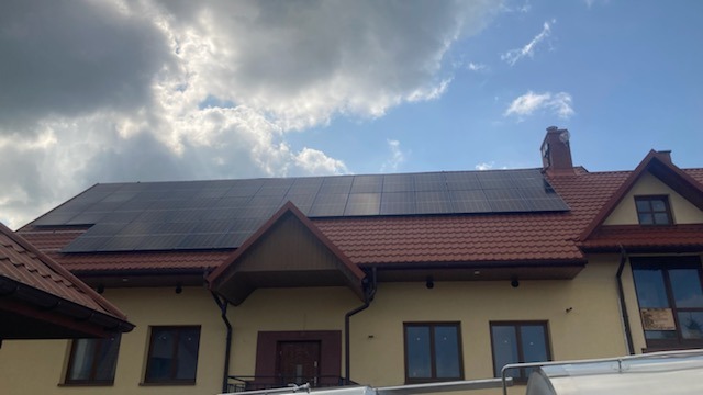 Panele fotowoltaiczne zamontowane na dachu dwupiętrowego domu z czerwoną dachówką, widoczne okna i komin. Częściowo pochmurne niebo w tle.
