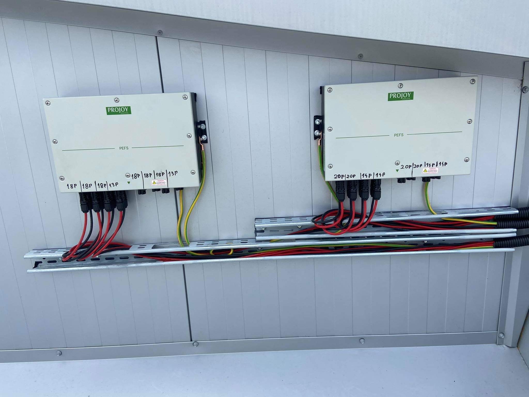 Dwa białe inwertery fotowoltaiczne marki Projoy, zamontowane na szarej ścianie, z podłączonymi przewodami i widoczną listwą montażową.