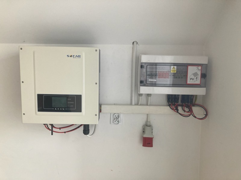 Biały inwerter SOFAR i skrzynka zabezpieczeń z symbolem PV zamontowane na białej ścianie, widoczne okablowanie i gniazdko elektryczne.