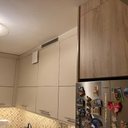 Meble Paweł Król - Fragment kuchni z beżowymi szafkami, brązową szafką wiszącą nad lodówką ozdobioną magnesami z podróży, oraz mozaiką nad blatem.