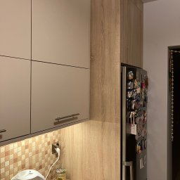 Meble Paweł Król - Kuchnia z beżowymi szafkami górnymi z podłużnymi uchwytami, drewnopodobnymi szafkami wysokimi obok lodówki ozdobionej magnesami, mozaiką nad blatem i czajnikiem elektrycznym Zelmer.