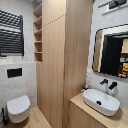 Meble Paweł Król - Nowoczesna łazienka z drewnianą zabudową: szafa z półkami, szafka pod umywalką z owalną białą umywalką nablatową i czarną armaturą, widok w lustrze na fragment kuchni.