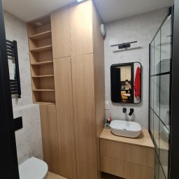 Meble Paweł Król - Nowoczesna łazienka z zabudową meblową z jasnego drewna, obejmującą szafę z półkami i szafkę pod umywalkę, z widocznym fragmentem kabiny prysznicowej w czarnej ramie i wiszącą toaletą.