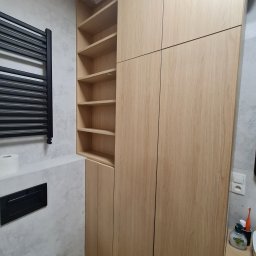 Meble Paweł Król - Wysoka, jasna szafa z drewna z otwartymi półkami w łazience, obok grzejnika drabinkowego i toalety.