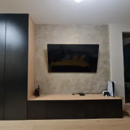 Czarno-beżowa zabudowa meblowa z telewizorem, konsolą PlayStation 5 i figurką słonia na blacie, na tle ściany z efektem dekoracyjnym.