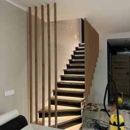 Meble Paweł Król - Nowoczesne schody drewniane z podświetleniem LED, pionowe lamele jako balustrada, widoczne narzędzia i odkurzacz na podłodze.