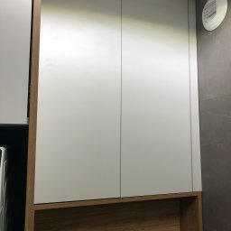 Meble Paweł Król - Biała szafa wnękowa z drewnianą półką na tle szarej ściany z wentylatorem.