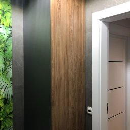 Meble Paweł Król - Wysoka, wąska szafa wnękowa z frontem z drewna i ciemnozielonym bokiem, wkomponowana w przestrzeń przy drzwiach i ścianie z szarego marmuru i zieloną tapetą z motywem roślinnym.