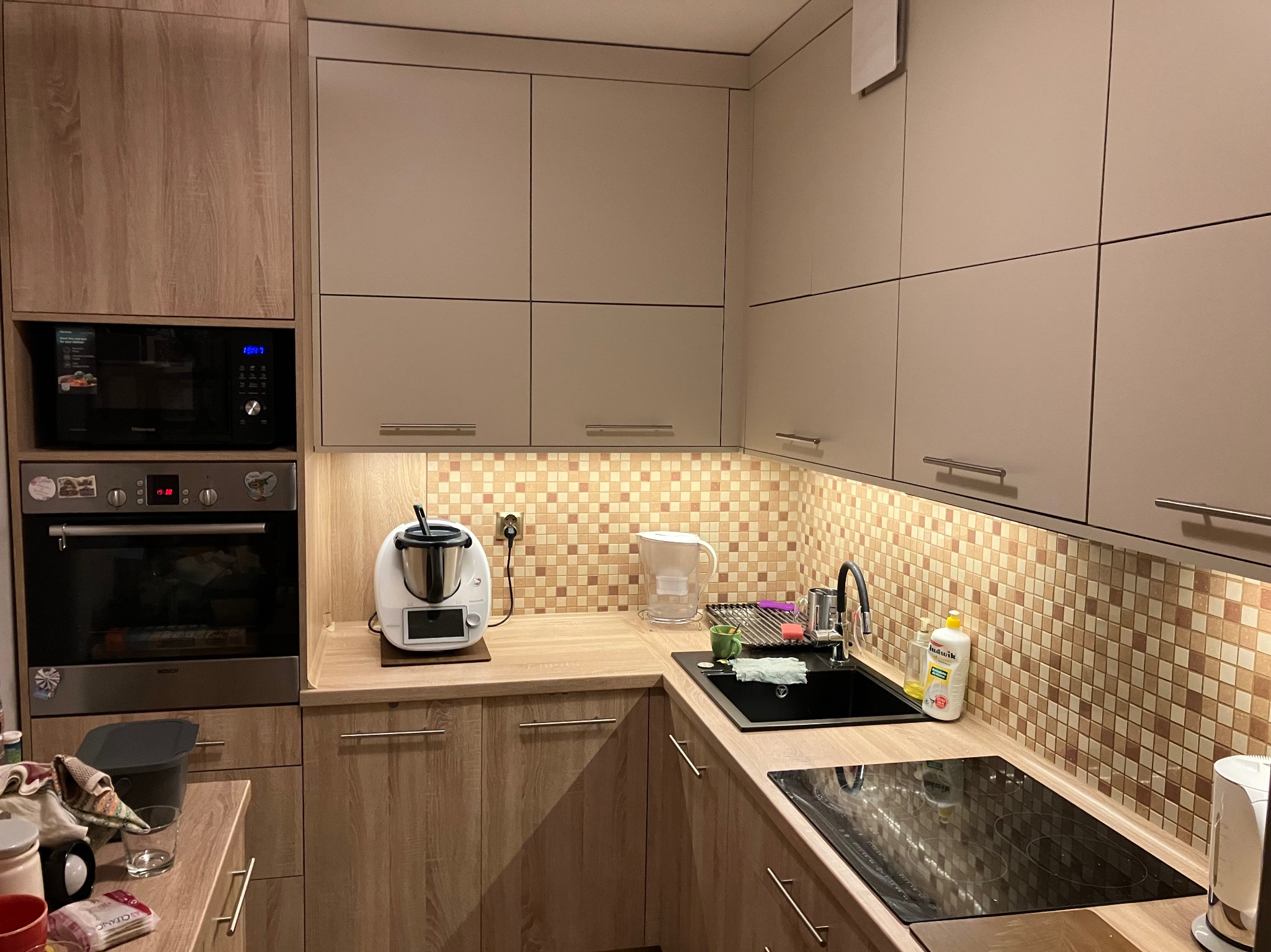 Jasna kuchnia z drewnianymi szafkami, mozaikową ścianą nad blatem, kuchenką indukcyjną i termomixem.