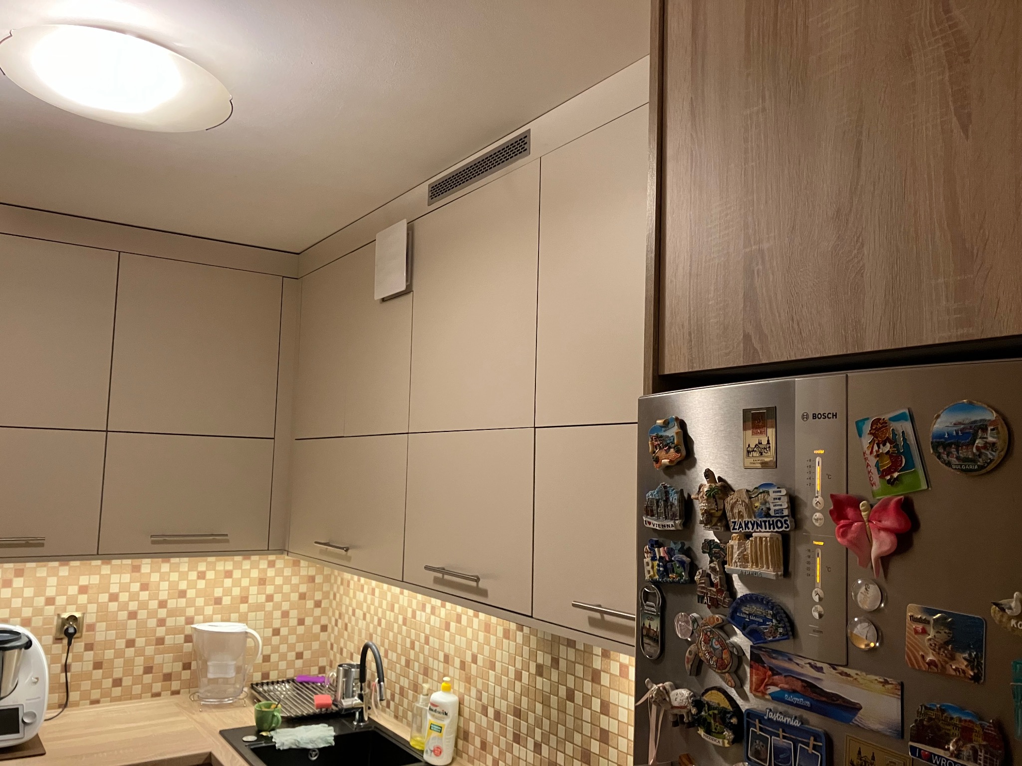 Jasna kuchnia z kremowymi szafkami górnymi, mozaikową ścianą nad blatem, czarnym zlewozmywakiem i lodówką Bosch ozdobioną magnesami z podróży.