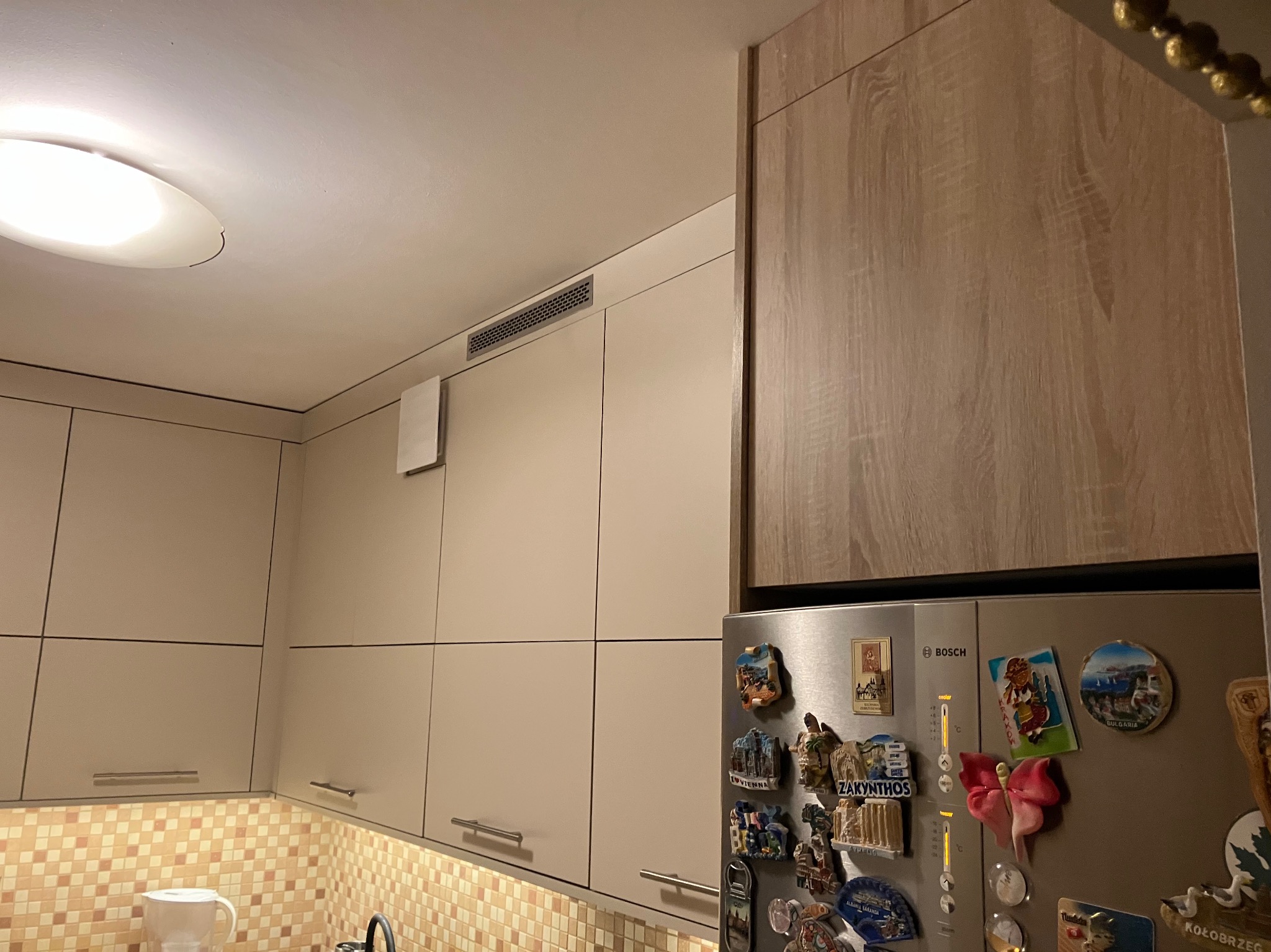 Fragment kuchni z beżowymi szafkami, brązową szafką wiszącą nad lodówką ozdobioną magnesami z podróży, oraz mozaiką nad blatem.