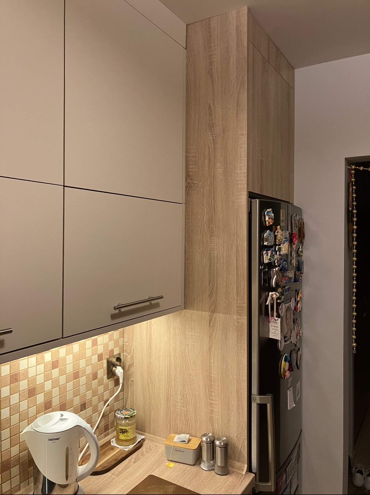 Kuchnia z beżowymi szafkami górnymi z podłużnymi uchwytami, drewnopodobnymi szafkami wysokimi obok lodówki ozdobionej magnesami, mozaiką nad blatem i czajnikiem elektrycznym Zelmer.