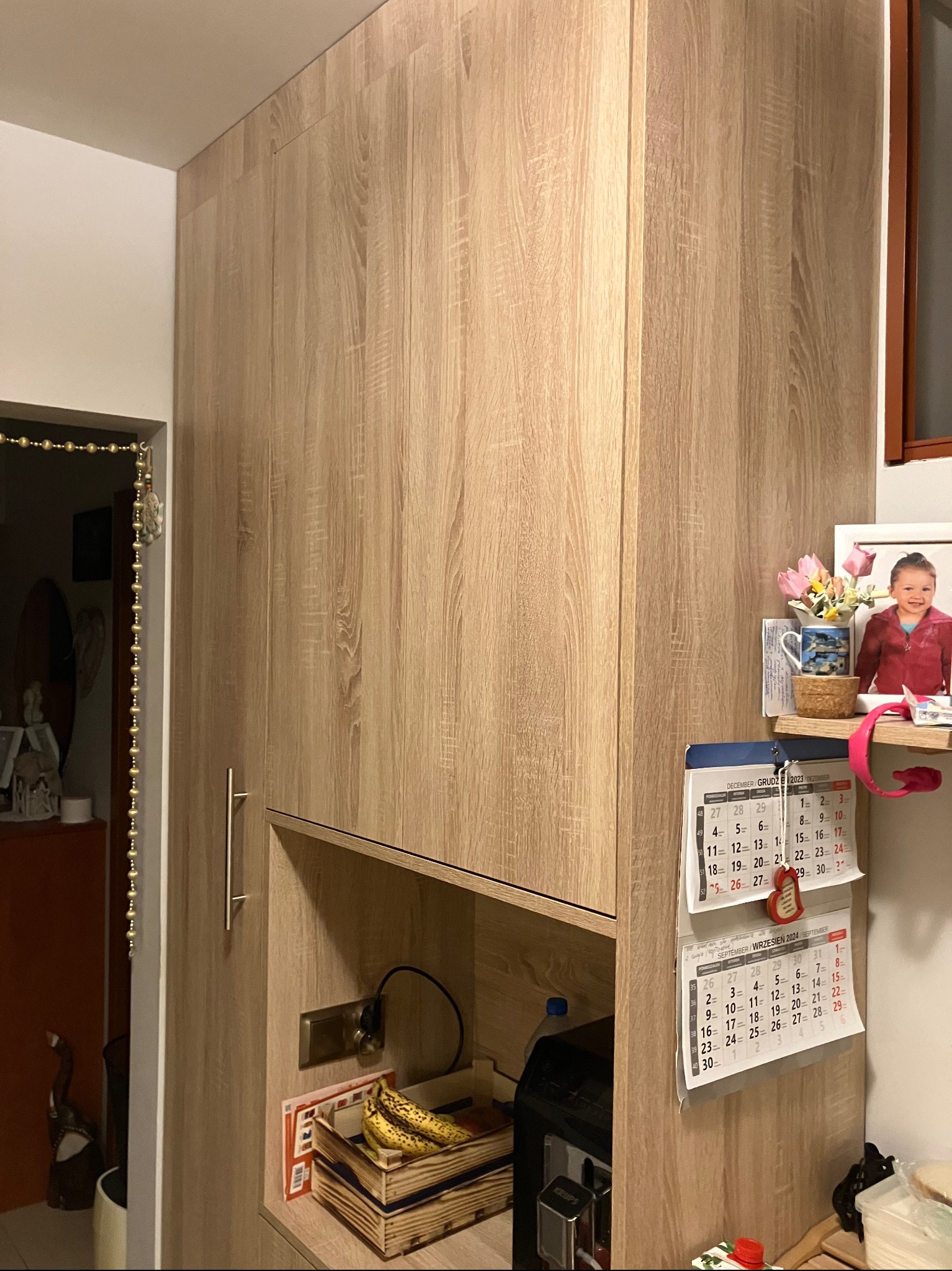 Wysoka szafa z jasnego drewna z widoczną fakturą słojów, z otwartą półką, na której leżą banany w drewnianej skrzynce, z kalendarzami i zdjęciem dziecka na półce obok okna.