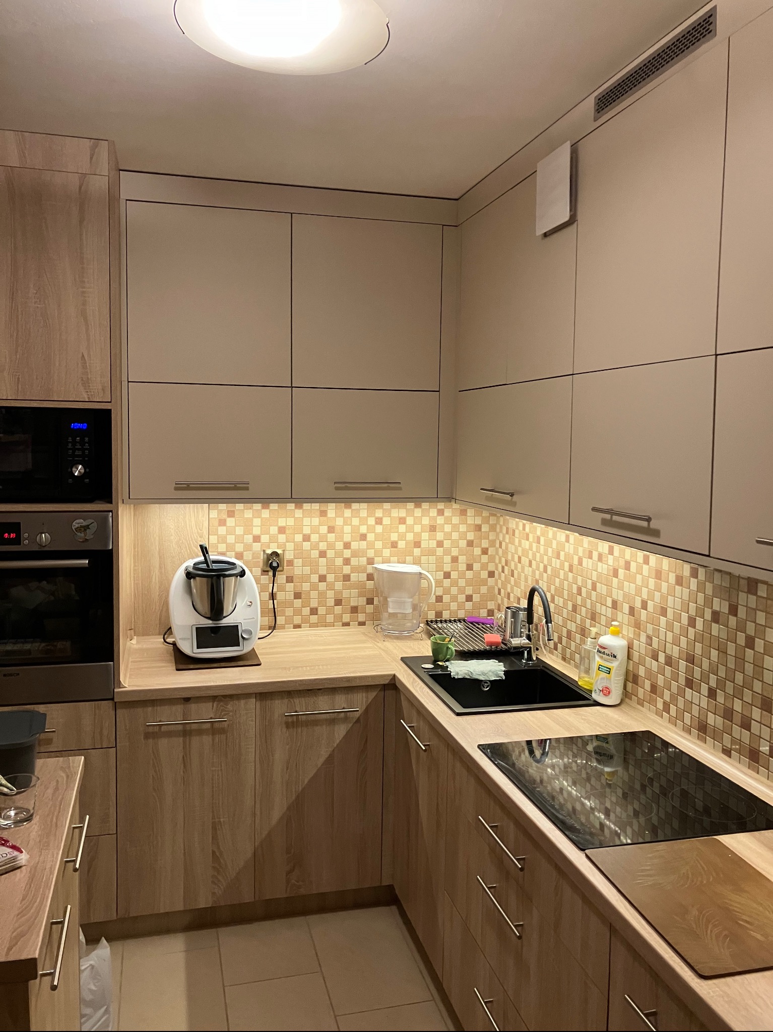 Nowoczesna kuchnia z drewnianymi i beżowymi frontami, mozaiką nad blatem, czarnym zlewem i płytą indukcyjną, wyposażona w sprzęt AGD, w tym Thermomix.