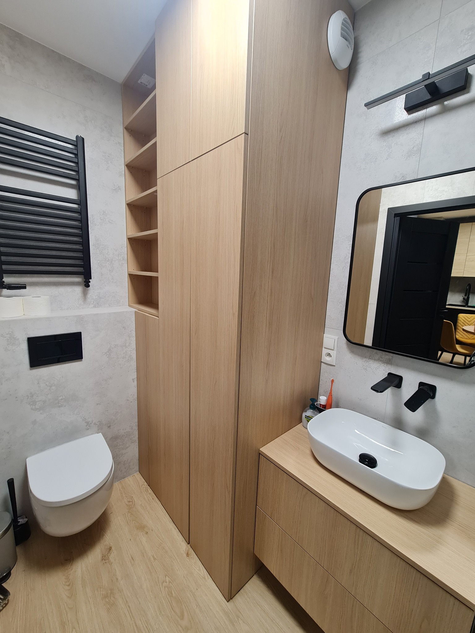 Nowoczesna łazienka z drewnianą zabudową: szafa z półkami, szafka pod umywalką z owalną białą umywalką nablatową i czarną armaturą, widok w lustrze na fragment kuchni.