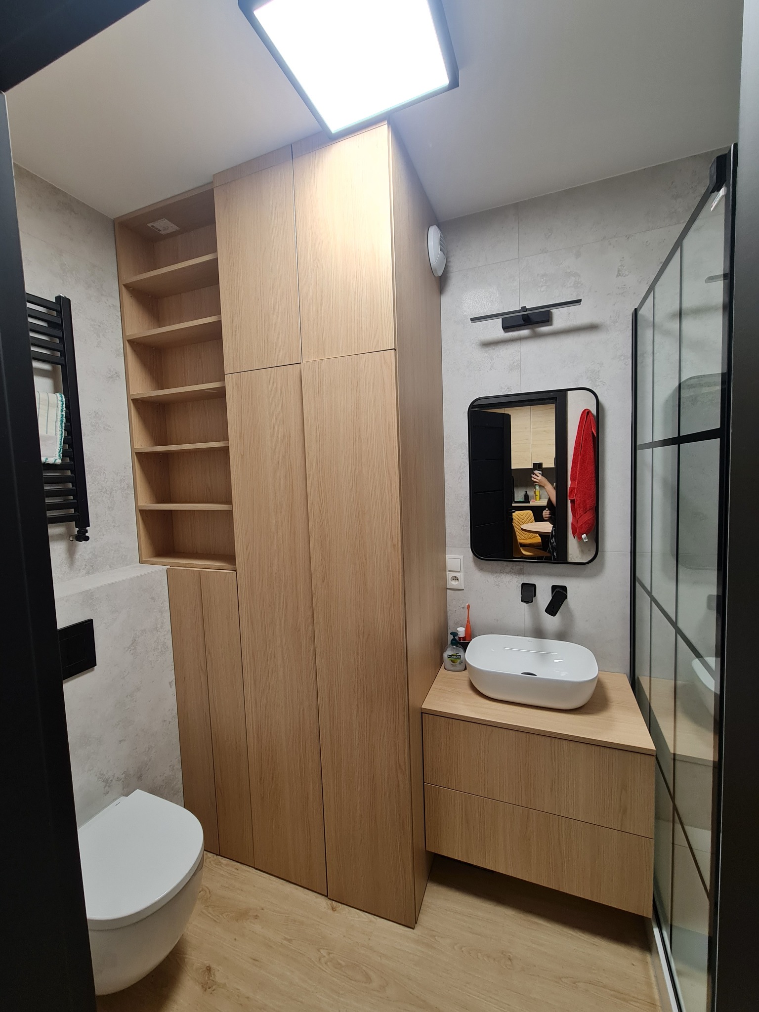 Nowoczesna łazienka z zabudową meblową z jasnego drewna, obejmującą szafę z półkami i szafkę pod umywalkę, z widocznym fragmentem kabiny prysznicowej w czarnej ramie i wiszącą toaletą.