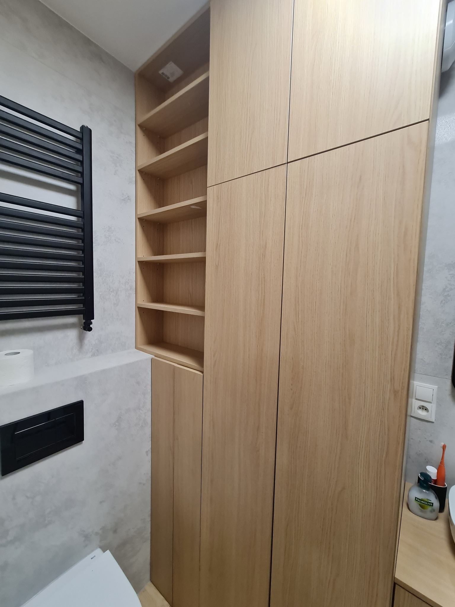 Wysoka, jasna szafa z drewna z otwartymi półkami w łazience, obok grzejnika drabinkowego i toalety.