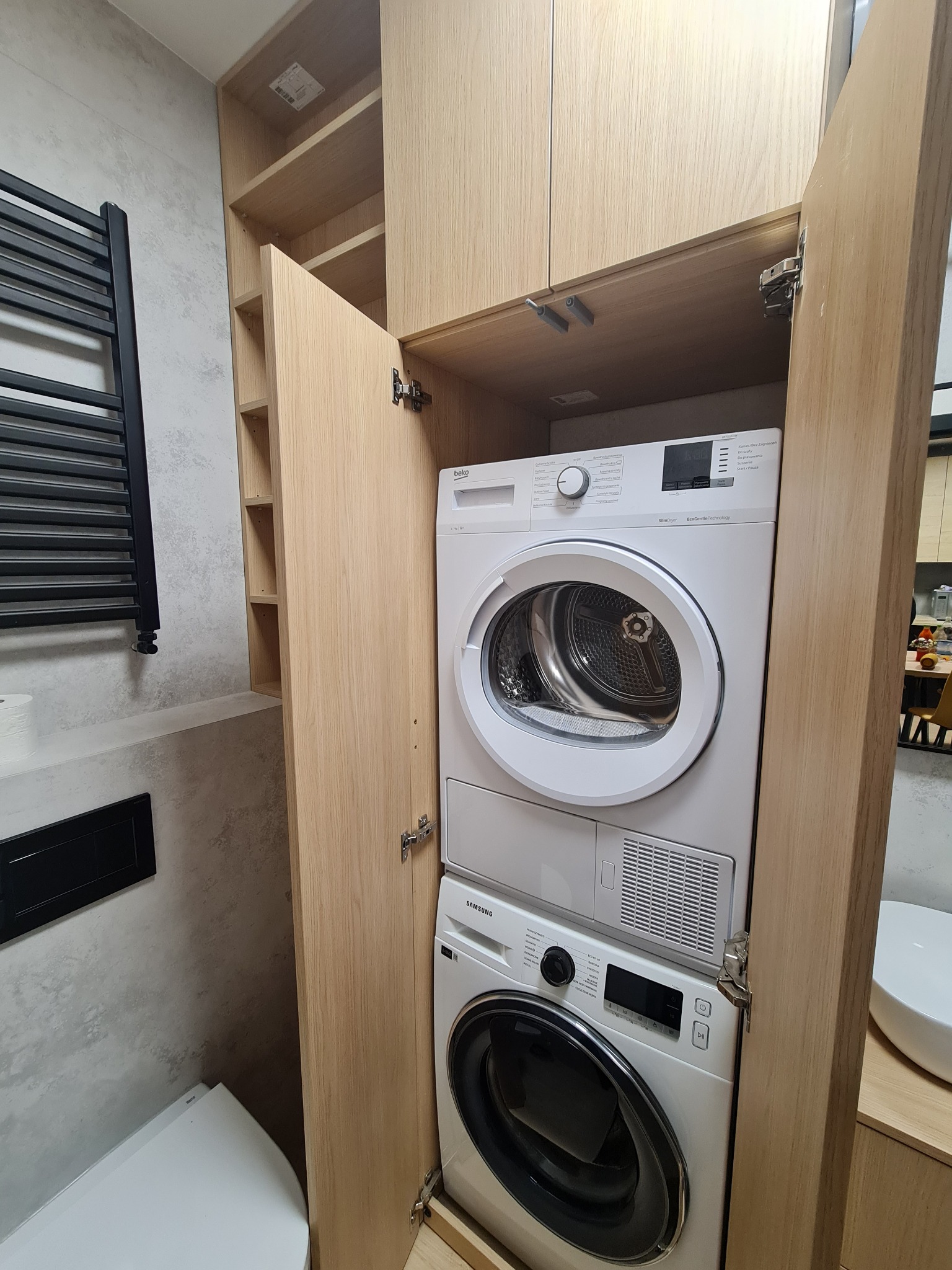 Pralka i suszarka w zabudowie z jasnego drewna w łazience, widoczne zawiasy i fragment toalety.