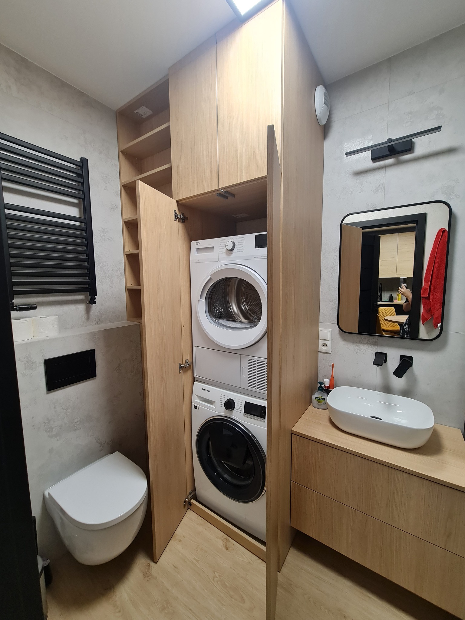 Nowoczesna łazienka z zabudowaną pralką i suszarką w szafie na wymiar z jasnego drewna, obok umywalka nablatowa i wisząca toaleta.