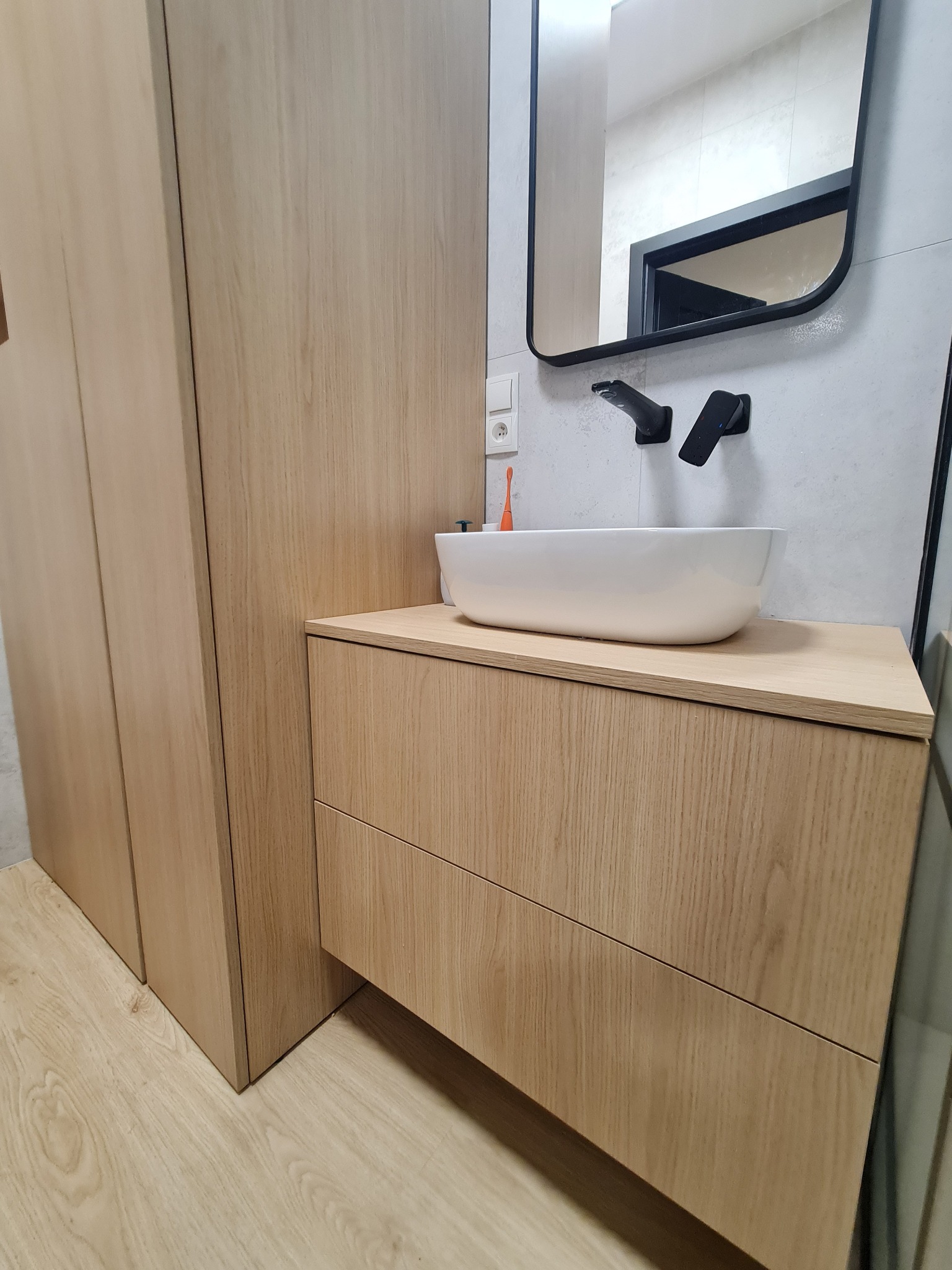 Nowoczesna łazienka z drewnianą szafką pod umywalkę nablatową, czarną armaturą i minimalistycznym lustrem w czarnej ramie.